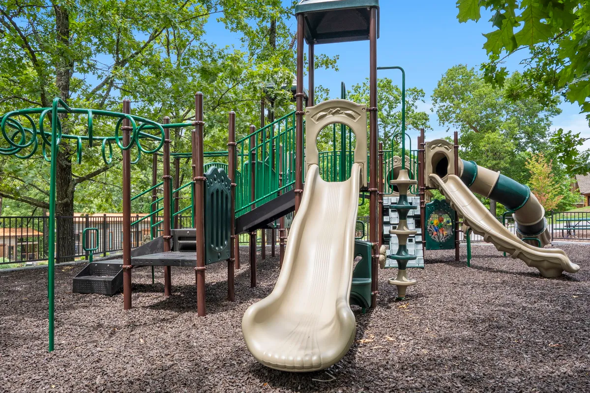 A colorful playground complete with slides, climbing structures, and a swing set sits at the heart of an outdoor gathering area. Children are seen playing while parents supervise and families picnic nearby, creating a scene full of energy and childhood joy.
