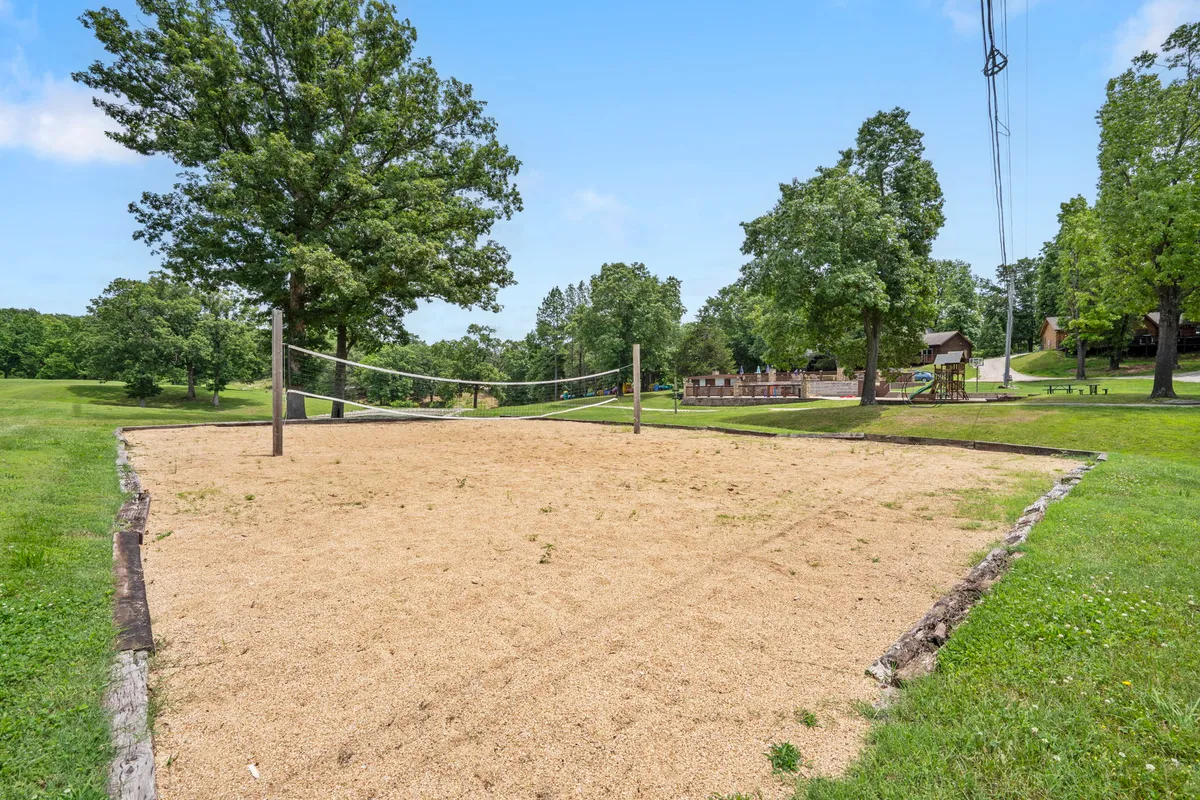 A sand volleyball court is surrounded by trees and distant resort buildings, with open sky above. The sandy area and surrounding greenery evoke a spirit of friendly competition and leisure within a natural setting.