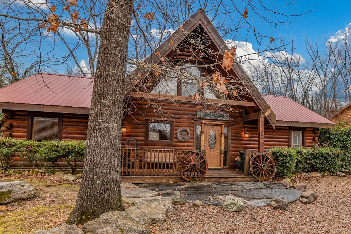 A welcoming log cabin exterior with a cozy front porch and wagon wheels, set along a stone pathway and surrounded by greenery. The porch is an inviting spot for relaxation and stargazing, embodying the warmth and tranquility of a woodland retreat.