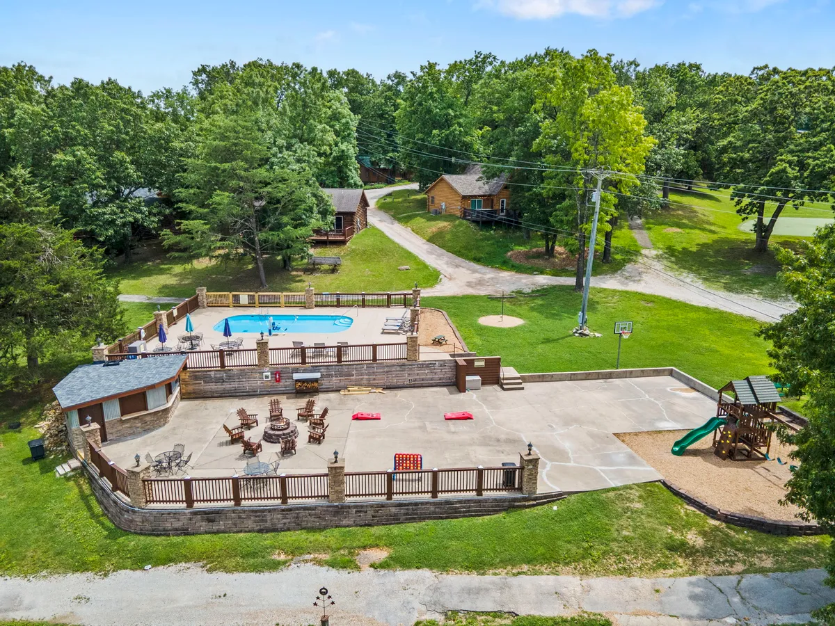 An exterior shot showcasing a swimming pool, patio area, playground, basketball hoop, and surrounding cabins. The atmosphere is lively yet relaxed, with spaces designed for swimming, sunbathing, barbecuing, and children's play—all surrounded by the community’s other cabins.