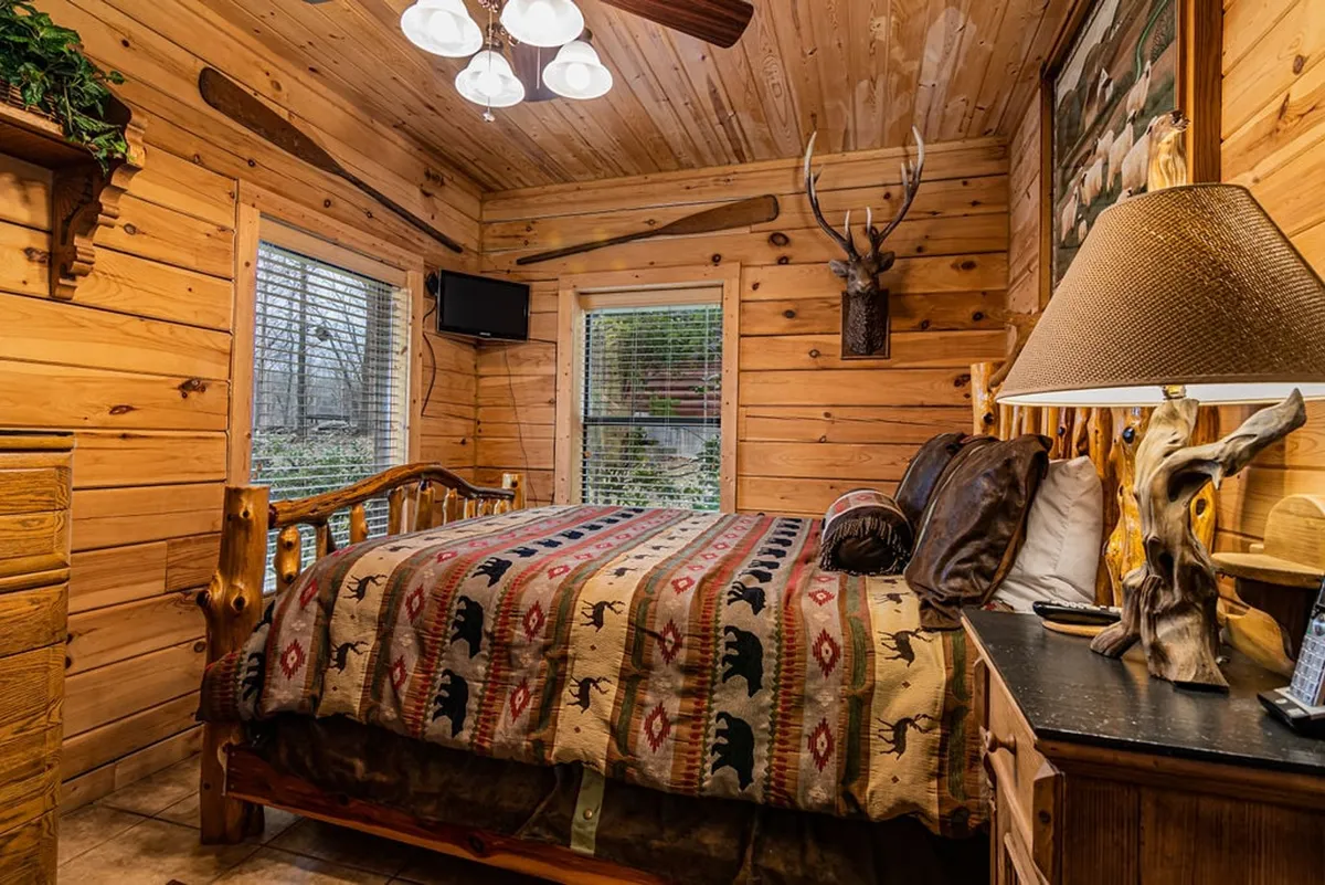 A warm and inviting bedroom featuring a wooden bed frame, colorful patterned bedding, wall-mounted antlers, a wooden dresser, and a nature-inspired lamp. The space feels both rustic and modern, with soft lighting creating a restful atmosphere.