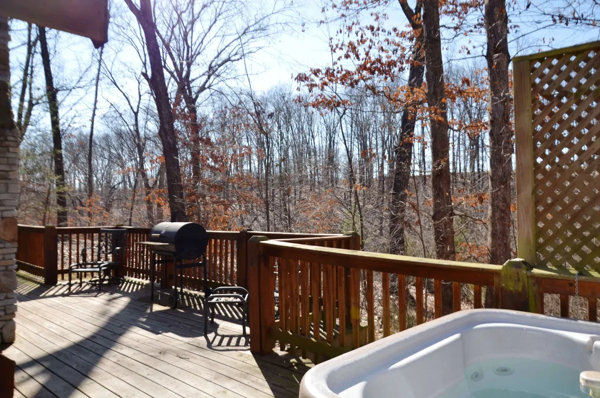 A wooden deck surrounded by a lush forest, featuring a hot tub, grill, and ample space for entertaining guests. The view highlights the connection to nature and the relaxing outdoor amenities.