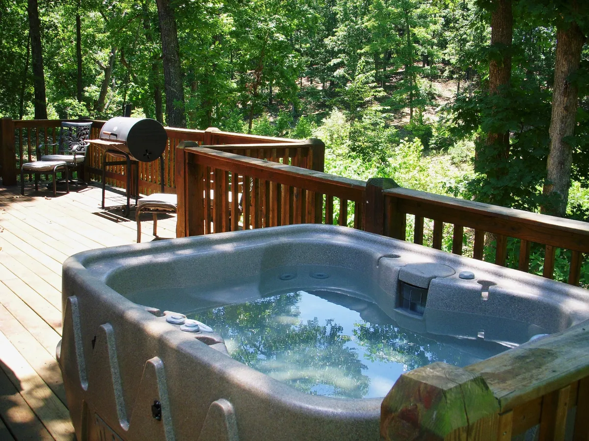 A spacious wooden deck bordered by trees, featuring a bubbling hot tub, a grill ready for outdoor cooking, and comfortable outdoor seating. The setting is tranquil, nestled beside the woods with soft evening sunlight filtering through the trees.