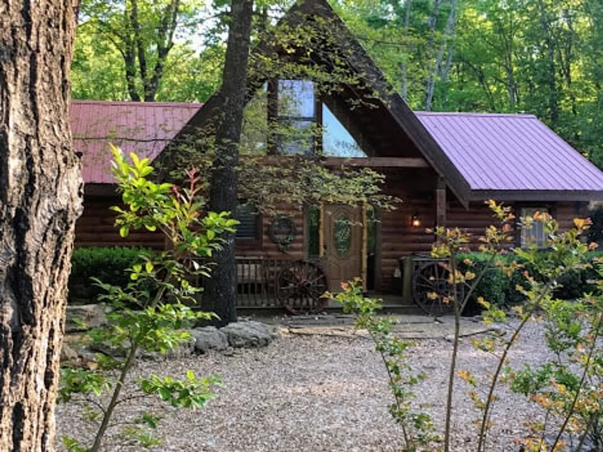 A charming log cabin stands amidst lush greenery, featuring a striking red metal roof and rustic wooden doors with glass panels. A gravel pathway leads up to the cabin, inviting guests to step into a serene, private world surrounded by the Ozark woodlands.