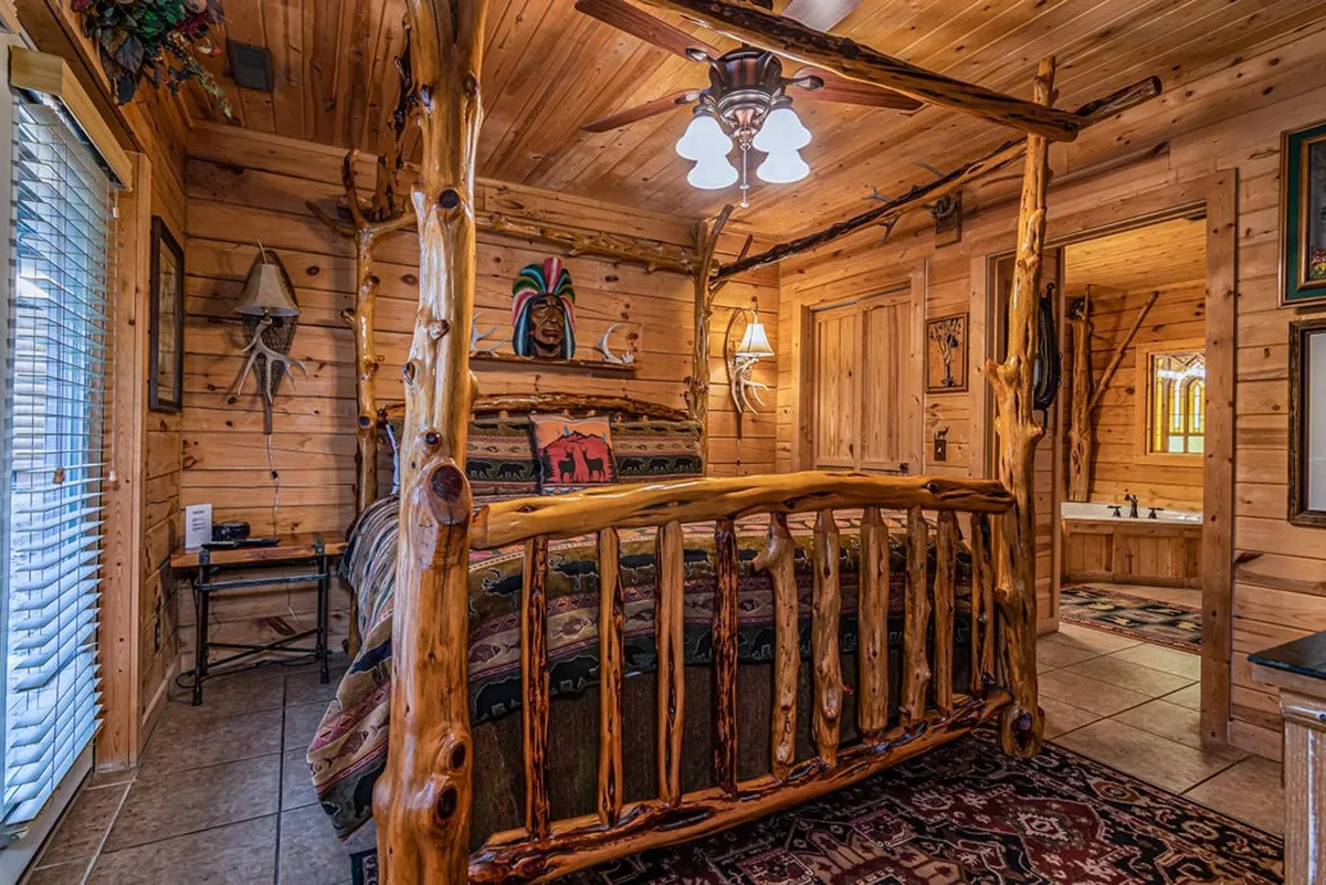 A cozy bedroom featuring a rustic log bed frame, decorative wall art, a wooden nightstand, and a warm light fixture. Natural sunlight streams in through a window, illuminating the wood finishes and inviting bedding, which exudes a blend of rustic elegance and comfort.