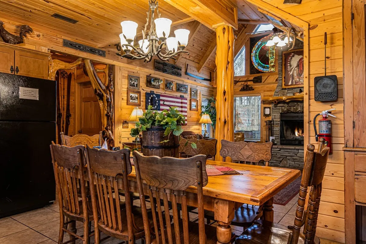 A spacious, warm dining area with a large wooden table set beneath a chandelier. The area features a stunning stone fireplace, white pine walls, stained glass window, and walls adorned with local artwork and decorative accents. The rustic setting is clearly designed for gatherings and shared meals, with cozy seating and welcoming ambiance.