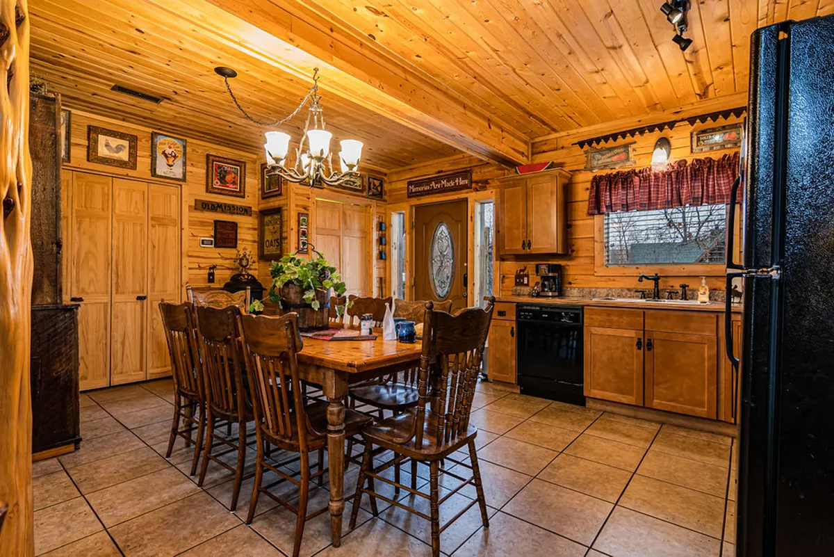 A wide-angle view of a rustic kitchen and dining space featuring a large wooden dining table, chandelier, kitchen cabinetry, and decorative wall art. The open-concept design allows glimpses of the adjoining kitchen, emphasizing togetherness and the flow of conversation from kitchen to table.